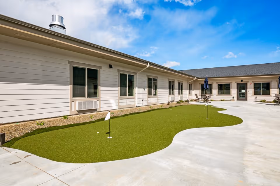 Courtyard with a small putting green, patio seating and a surrounding single-story senior living building under a blue sky.