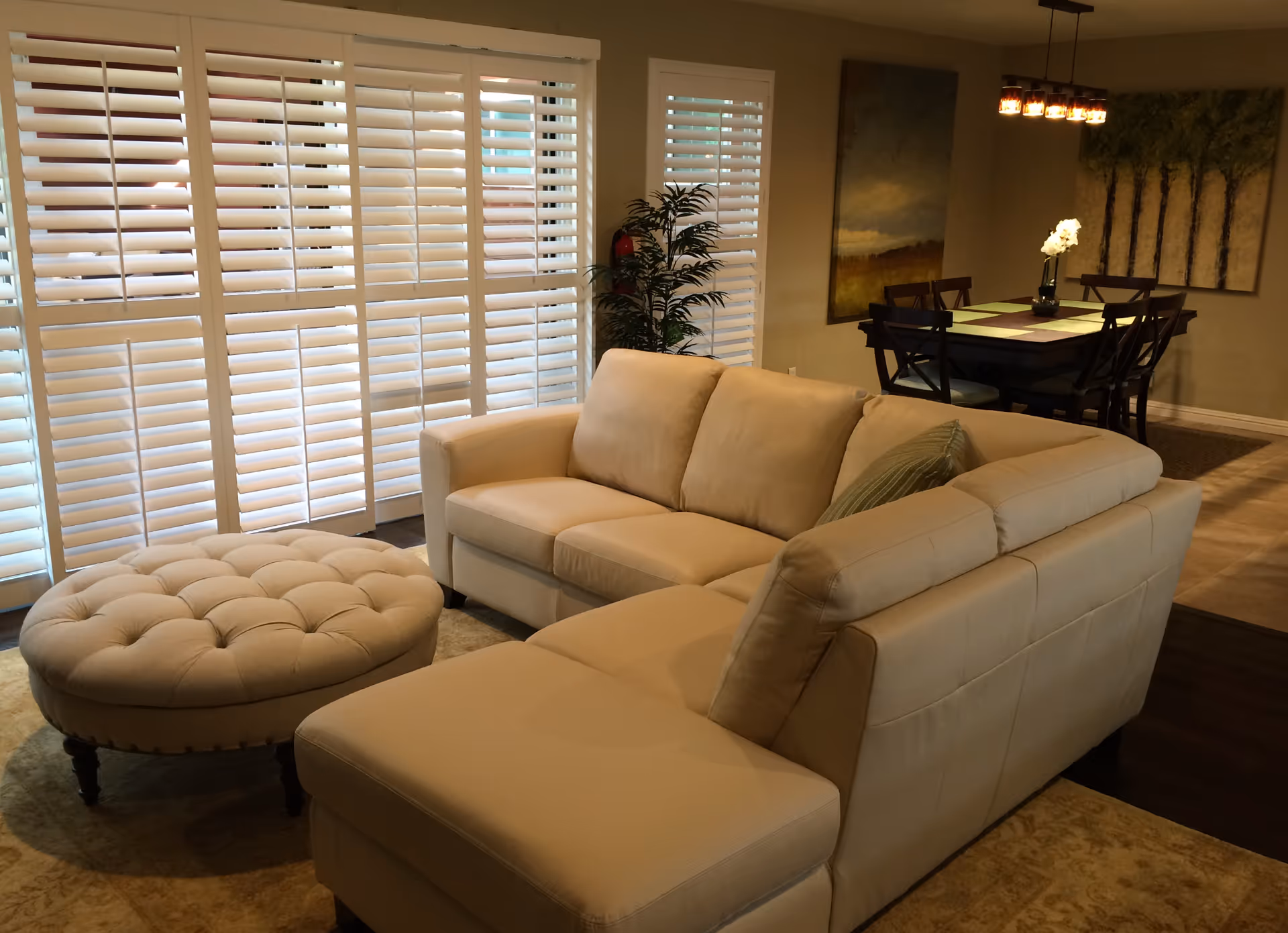 A cozy living room area with a beige sectional sofa and a round tufted ottoman. Behind the sofa, there are large white plantation shutters covering the windows. In the background, there is a dining area with a dark wooden table, six chairs, a vase with white flowers, and two large paintings on the wall. A modern chandelier with multiple lights hangs above the dining table.