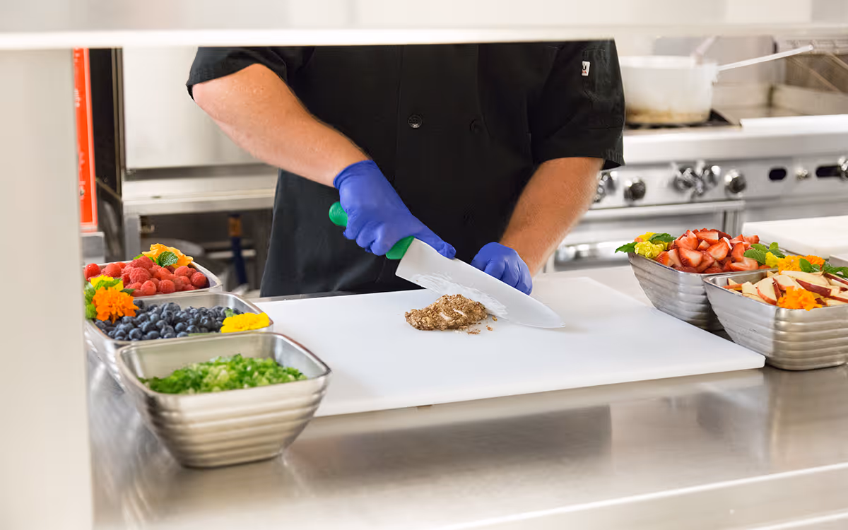 A person wearing blue gloves and a black chef coat is chopping food on a white cutting board in a commercial kitchen. Surrounding the cutting board are metal trays filled with fresh fruits and vegetables including raspberries, blueberries, strawberries, apples, and chopped green onions.