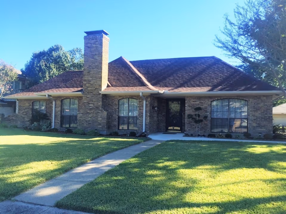 Single-story brick house with a large chimney, three arched windows, a black front door, and a well-maintained green lawn with a concrete walkway leading to the entrance. Trees and shrubs surround the house under a clear blue sky.