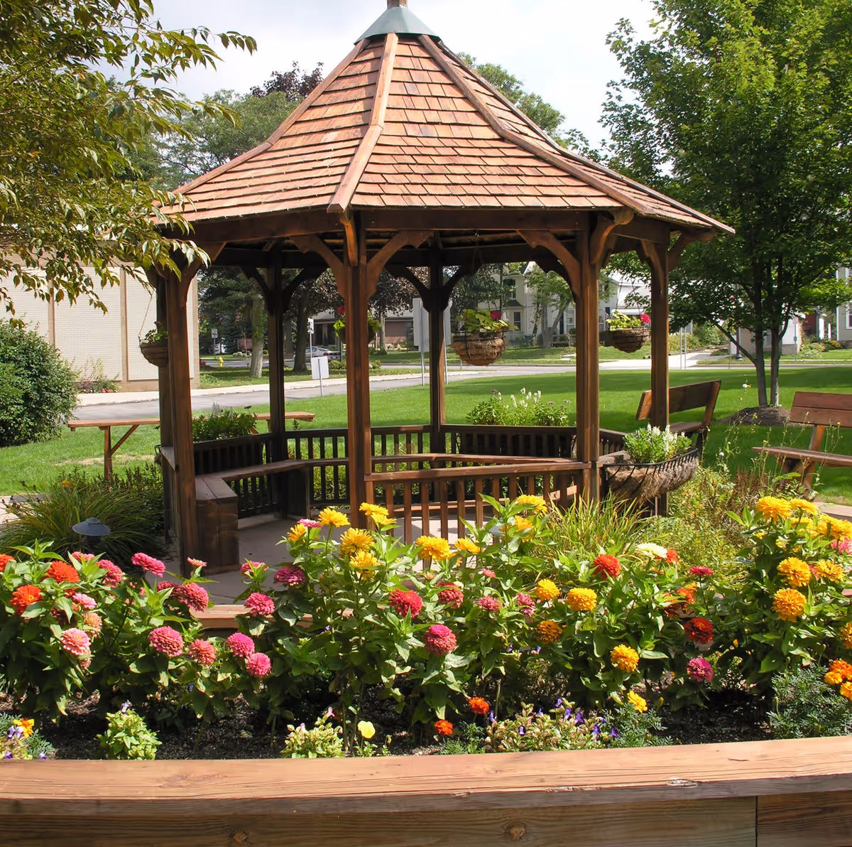 A wooden gazebo with a shingled roof surrounded by colorful flowers and greenery in a garden area. There are benches inside the gazebo and additional benches nearby. Trees and a street with houses are visible in the background.