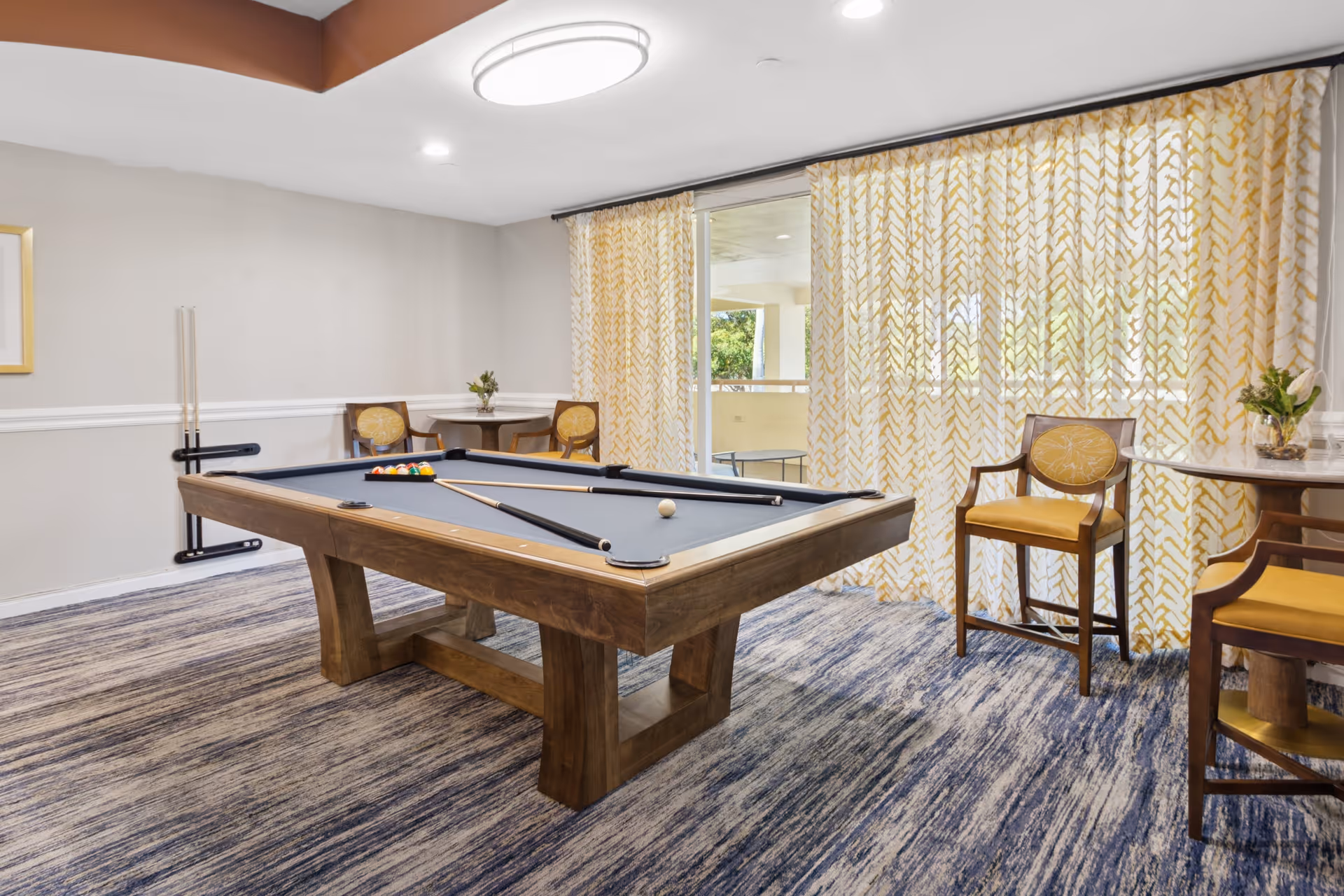 A well-lit game room with a wooden pool table set up for a game, featuring a blue felt surface and pool cues resting on the table. The room has patterned carpet flooring, two small round tables with yellow cushioned chairs, and large windows covered with yellow and white patterned curtains allowing natural light to enter.