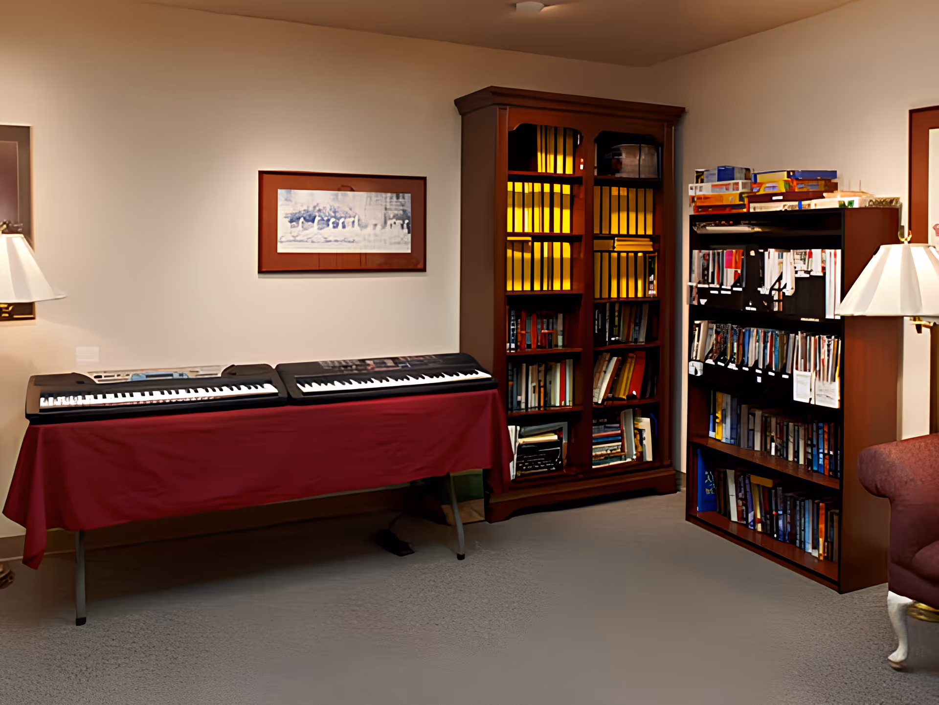 A cozy room with two electronic keyboards on a table covered with a red cloth, a wooden bookshelf filled with books and binders, another wooden bookshelf with various books and board games on top, a framed picture on the wall, and a floor lamp next to a red upholstered chair.