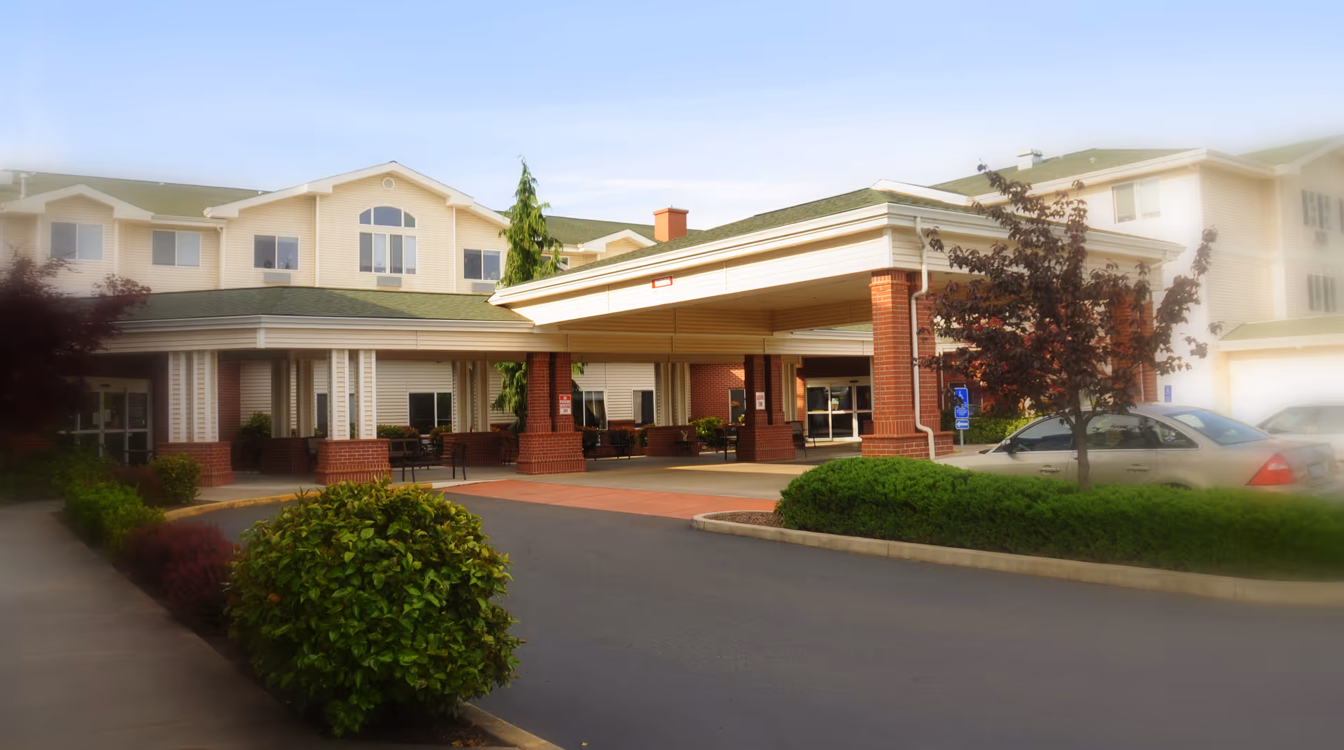 Exterior view of Avamere at Three Fountains senior living facility showing the main entrance with a covered drop-off area, surrounded by greenery and parked cars.