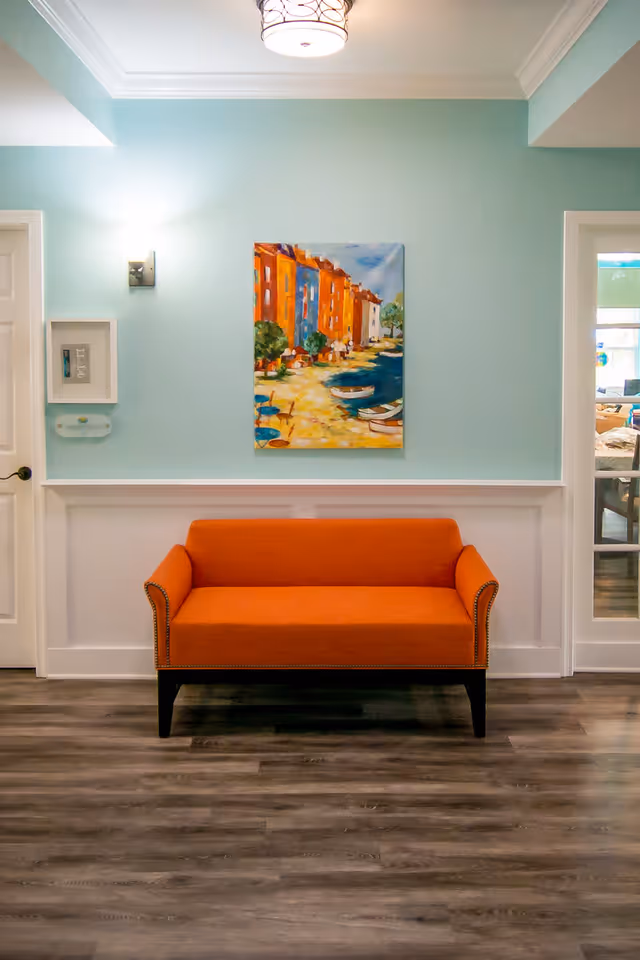 An interior room with a bright orange upholstered bench against a light blue wall. Above the bench is a colorful painting depicting a row of buildings and boats by the water. The room has wood flooring, white trim, a ceiling light fixture, and two doors on either side of the bench.