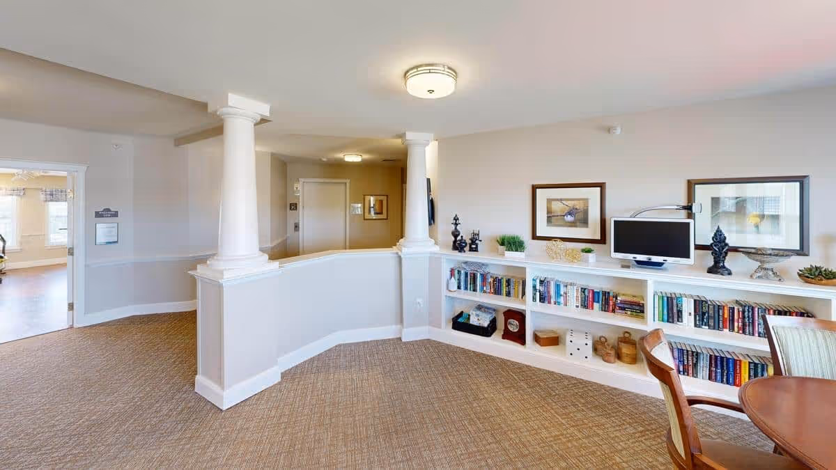 Interior view of a senior living facility common area with beige walls and carpeted floor. The space features two white columns on a half wall, built-in white bookshelves filled with books and decorative items, framed artwork on the wall, a small flat-screen TV, and a round wooden table with chairs. There is a doorway leading to another room with large windows and natural light.
