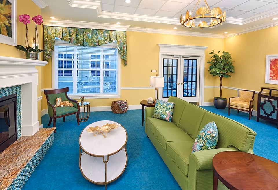 A bright and cozy living room with yellow walls, blue carpet, and white crown molding. The room features a green sofa with patterned cushions, a white oval coffee table with a decorative centerpiece, a green armchair with a small stuffed animal, a side table with a lamp, a potted plant, and a fireplace with a tiled surround. Large windows with floral valances and French doors lead to an outdoor area.