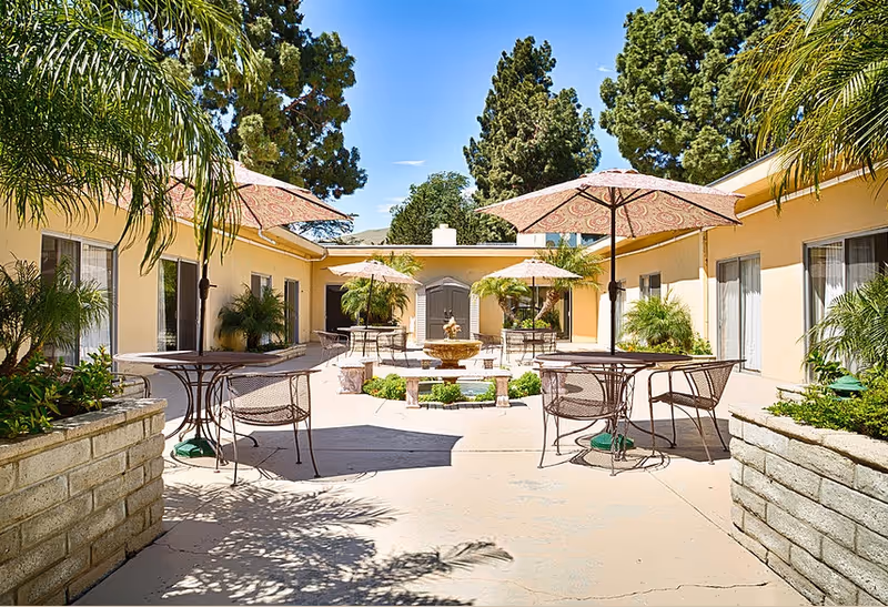 Outdoor courtyard area at Ventura Post Acute with round metal tables and chairs under patterned umbrellas, surrounded by beige buildings with windows, potted plants, palm trees, and a central water fountain. Tall trees are visible in the background under a clear blue sky.