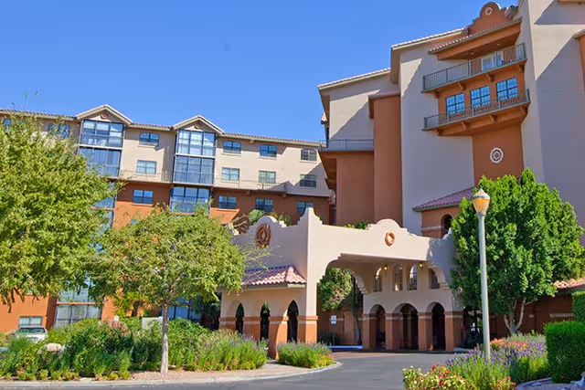 Exterior view of Casa del Sol, a multi-story building with a southwestern architectural style featuring stucco walls, arched entryways, and balconies. The foreground includes landscaped greenery, trees, and a driveway under a clear blue sky.
