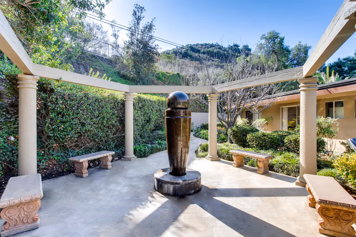 Outdoor courtyard area with a central dark stone sculpture surrounded by four beige stone benches. The courtyard is framed by beige columns supporting a partial pergola structure. Green bushes and trees surround the area, with a hillside and clear blue sky in the background.