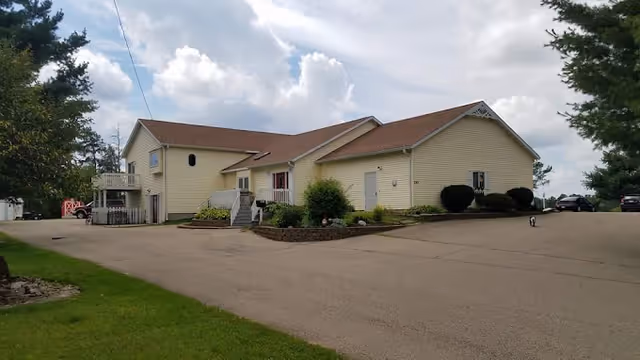 Exterior front view of a single-story yellow residential building with a paved driveway, landscaped shrubs, and a cloudy sky.