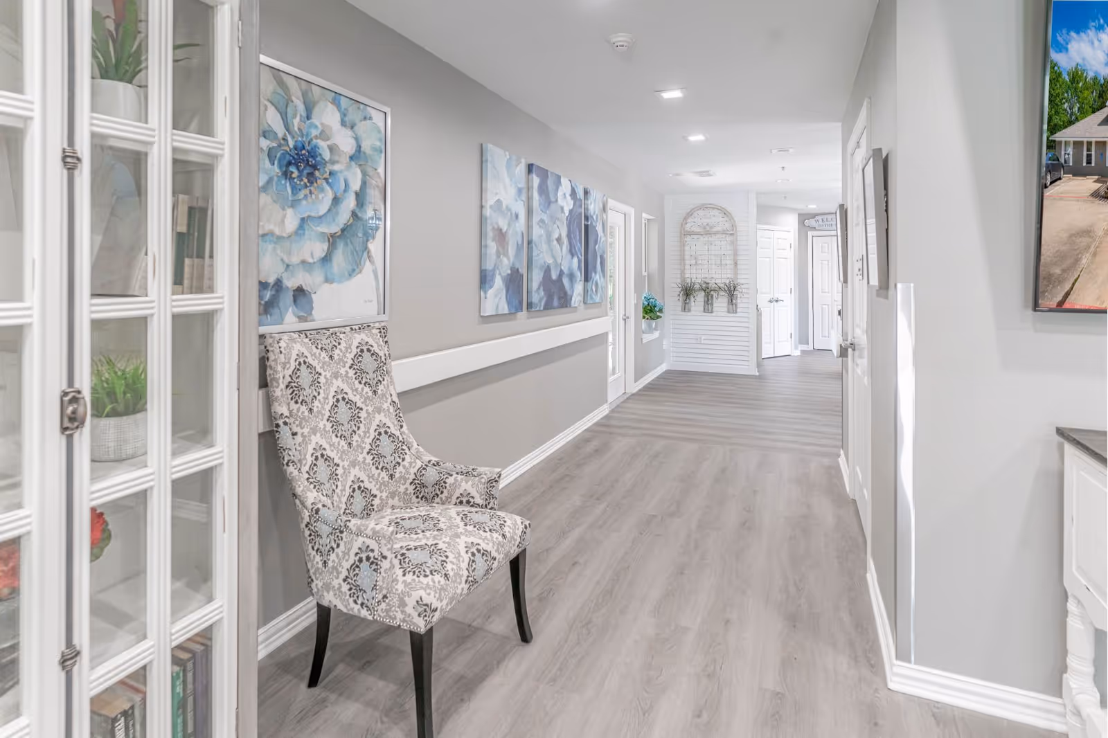 A bright, clean hallway in an assisted living facility with light gray walls and wood flooring. There is a patterned upholstered chair against the left wall, decorative blue floral artwork hanging above it, and a glass-front cabinet with plants and books inside. The hallway extends to white doors and a decorative wall piece with plants at the far end.