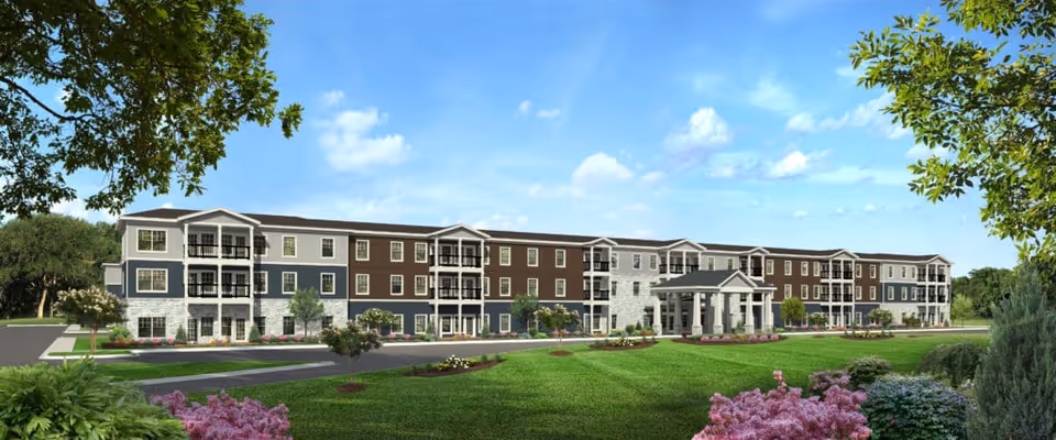 Exterior view of a large, three-story senior living facility building with a mix of gray, brown, and white siding, multiple balconies, and a covered entrance. The building is surrounded by well-maintained green lawns, landscaped flower beds, and trees under a blue sky with some clouds.