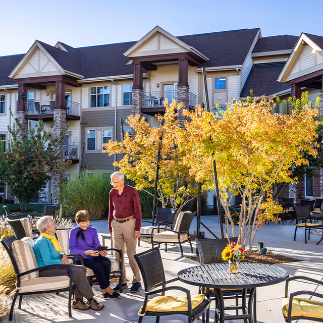 Three elderly people sitting and standing around outdoor patio chairs and tables in front of a multi-story residential building with autumn trees and clear blue sky.