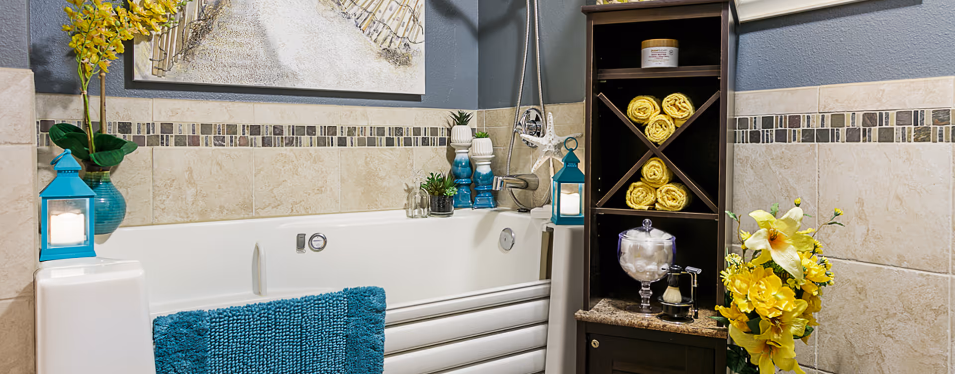 A bathroom featuring a white bathtub with a blue textured bath mat hanging over the side. The walls are tiled with beige tiles and a decorative mosaic border. There are blue lanterns with candles on either side of the tub, a vase with yellow flowers, rolled yellow towels stored in a dark wooden shelving unit, and small decorative plants and items on the tub edge and shelf. A piece of wall art hangs above the bathtub.