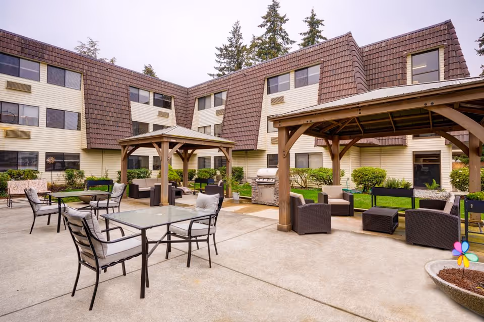 Outdoor courtyard area of a senior living facility with multiple seating arrangements including glass-top tables with cushioned chairs and wicker lounge chairs under wooden pergolas. The building surrounding the courtyard has beige siding and brown shingled mansard roofs. There are some shrubs and trees around the perimeter and a colorful pinwheel decoration in a planter.