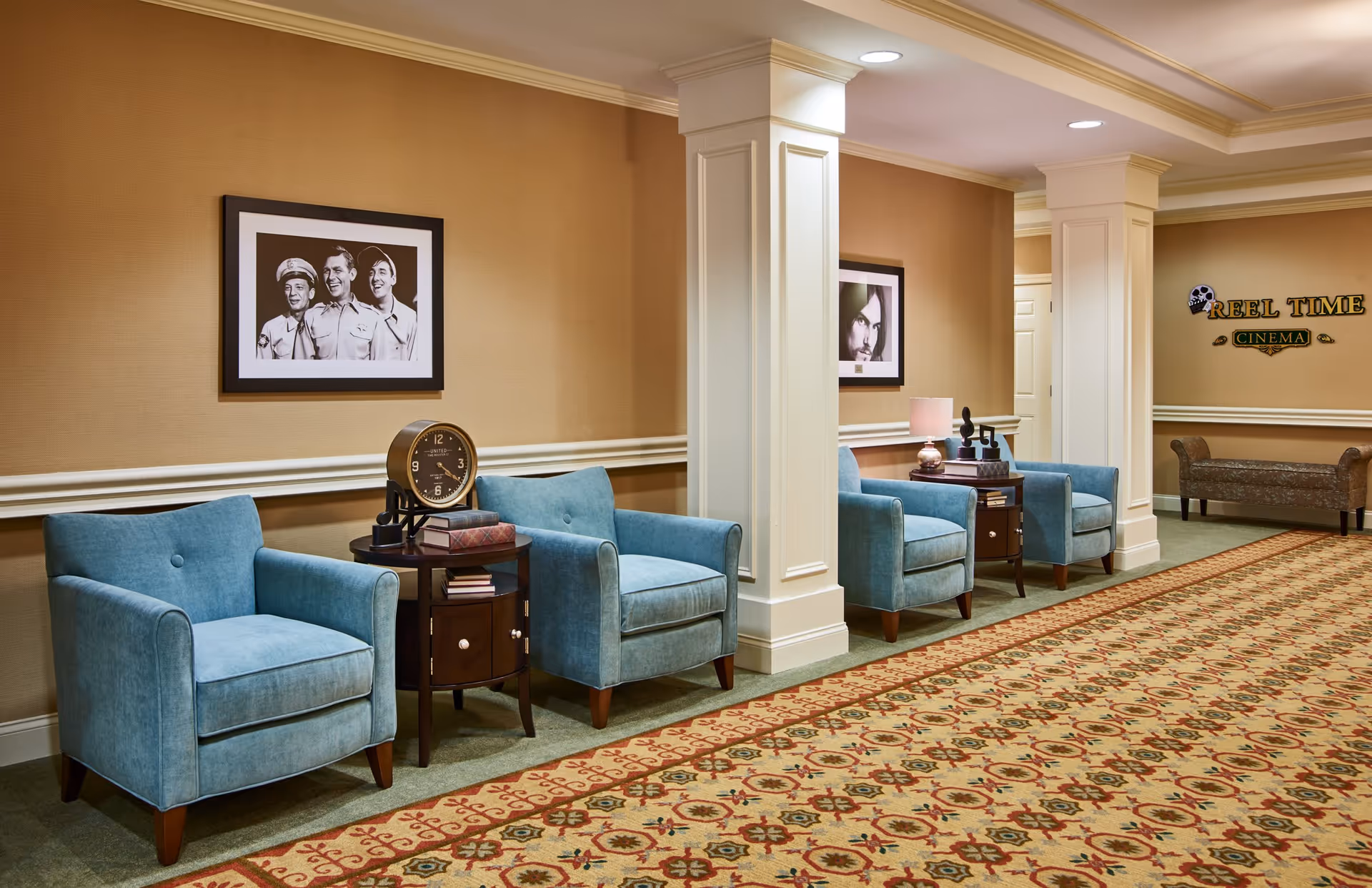 A hallway seating area with four blue upholstered armchairs arranged in pairs along a beige wall. Between each pair is a dark wooden side table with decorative items including a clock, books, and a lamp. The wall features framed black and white photographs, and there are white columns dividing the space. A patterned carpet covers the floor, and a sign on the far wall reads 'REEL TIME CINEMA' with a skull graphic.
