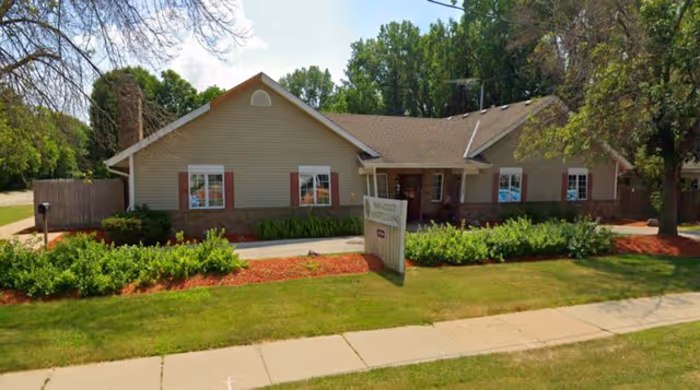 Front exterior of a single-story beige building with a lawn, shrubs, and a small sign by the sidewalk.