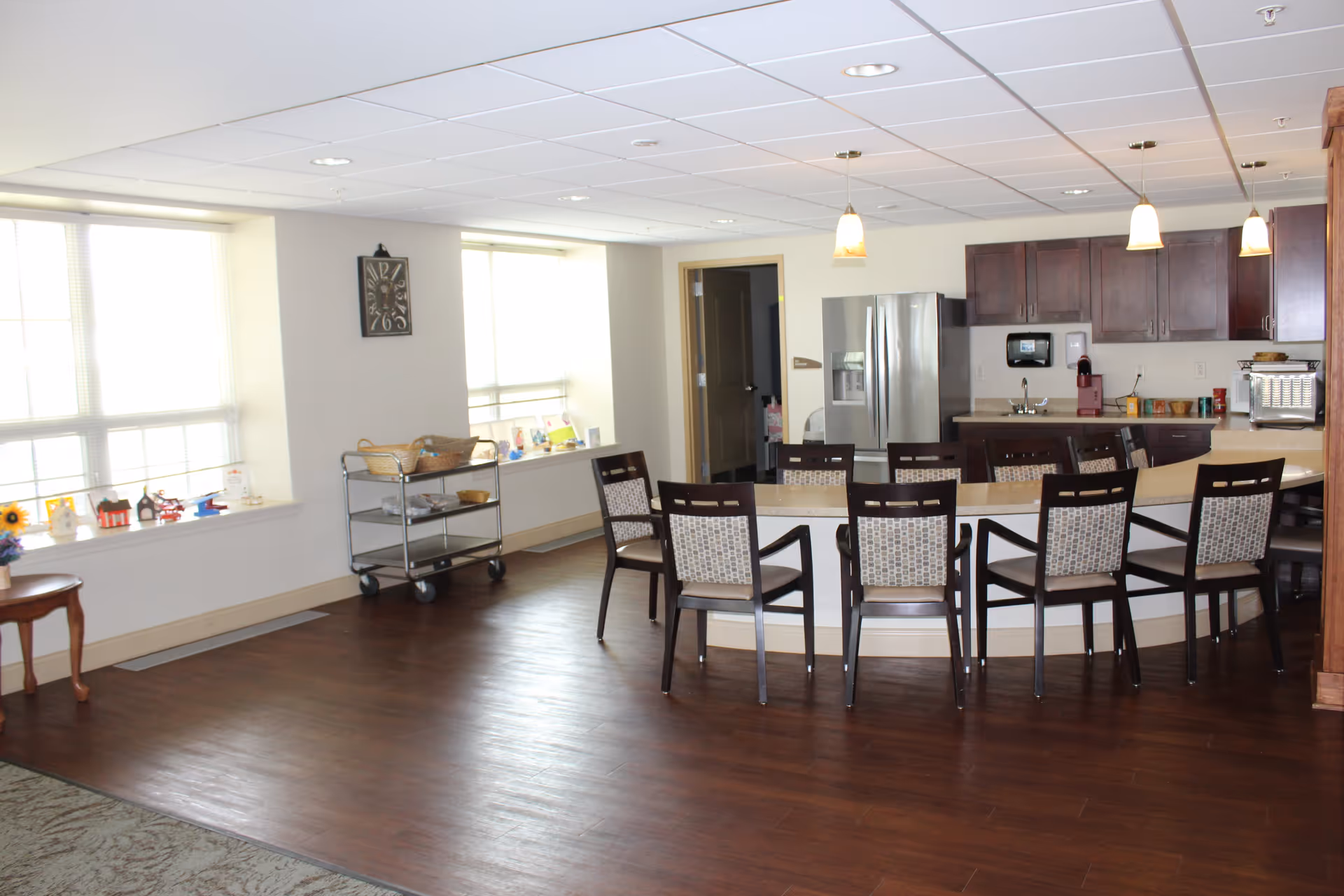A bright and spacious senior living facility kitchen and dining area with a curved counter surrounded by eight chairs. The kitchen features dark wood cabinets, a stainless steel refrigerator, a coffee machine, and a microwave. Large windows allow natural light to fill the room, and a clock hangs on the wall between the windows. A small cart with baskets and items is positioned near the windows.