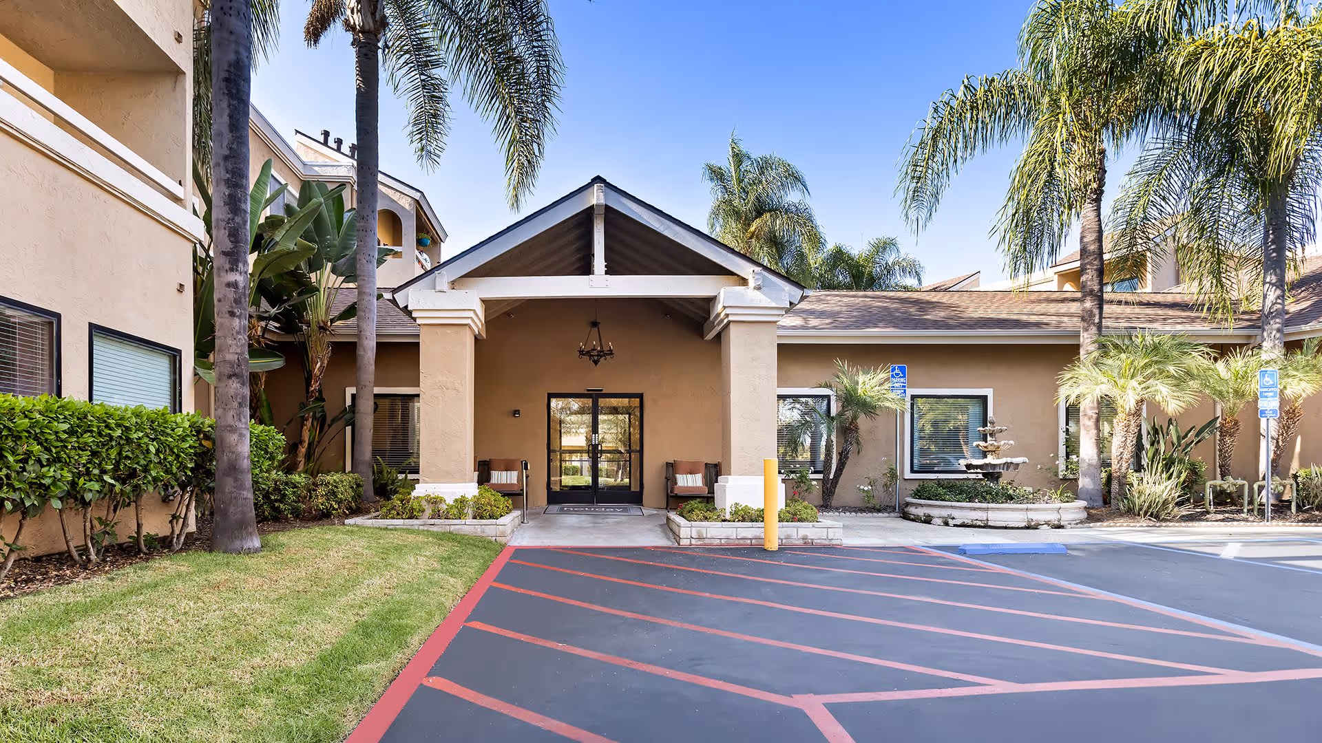 Entrance of a senior living facility with a covered porch supported by columns, two benches on either side of the door, palm trees, manicured bushes, a fountain, and a parking area with handicap parking spaces.