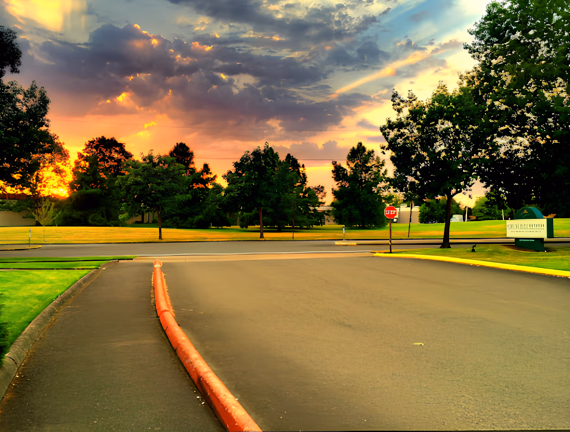 A paved road with a red curb leading to a stop sign at an intersection, surrounded by green grass and trees under a dramatic sunset sky with clouds.