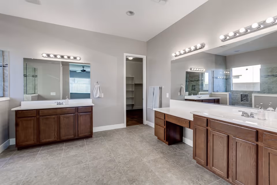 A spacious bathroom with two separate wooden vanities, each with a large mirror and bright vanity lights above. The room has light gray walls, tiled flooring, and a doorway leading to a walk-in closet. A window near one vanity lets in natural light.
