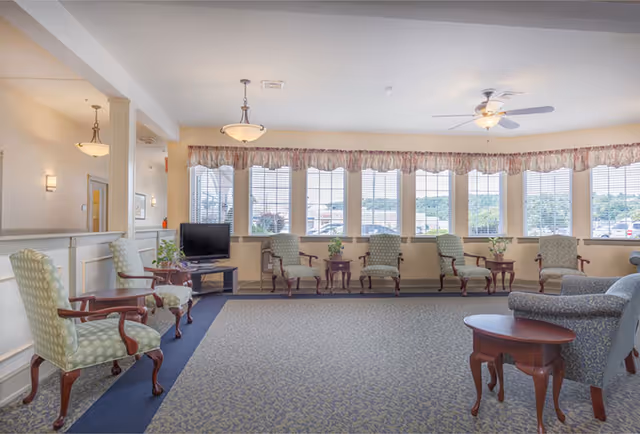 A spacious and well-lit common area with multiple upholstered armchairs arranged along the walls and around small wooden tables. Large windows with blinds and valances let in natural light, and a ceiling fan with a light fixture is mounted on the ceiling. A small TV is placed on a stand in the corner near the windows.