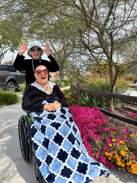 Two elderly women outdoors on a sunny day. One woman is sitting in a wheelchair covered with a blue and white patterned blanket, smiling and wearing sunglasses. The other woman is standing behind her, wearing a hat and sunglasses, raising her hands in a joyful gesture. They are on a sidewalk next to a garden bed with vibrant pink and orange flowers and trees in the background.