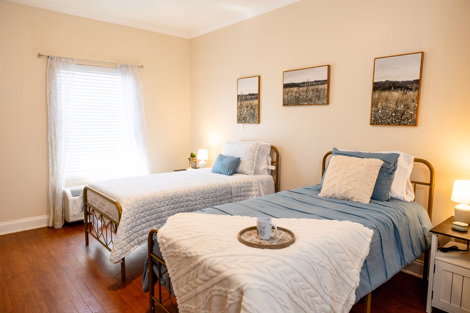 A cozy bedroom with two single beds, one with a blue bedspread and the other with a light gray quilt. Each bed has pillows and a small nightstand with a lamp beside it. Three framed pictures of a field hang on the wall above the beds. A window with sheer curtains lets in natural light, and the floor is wooden.
