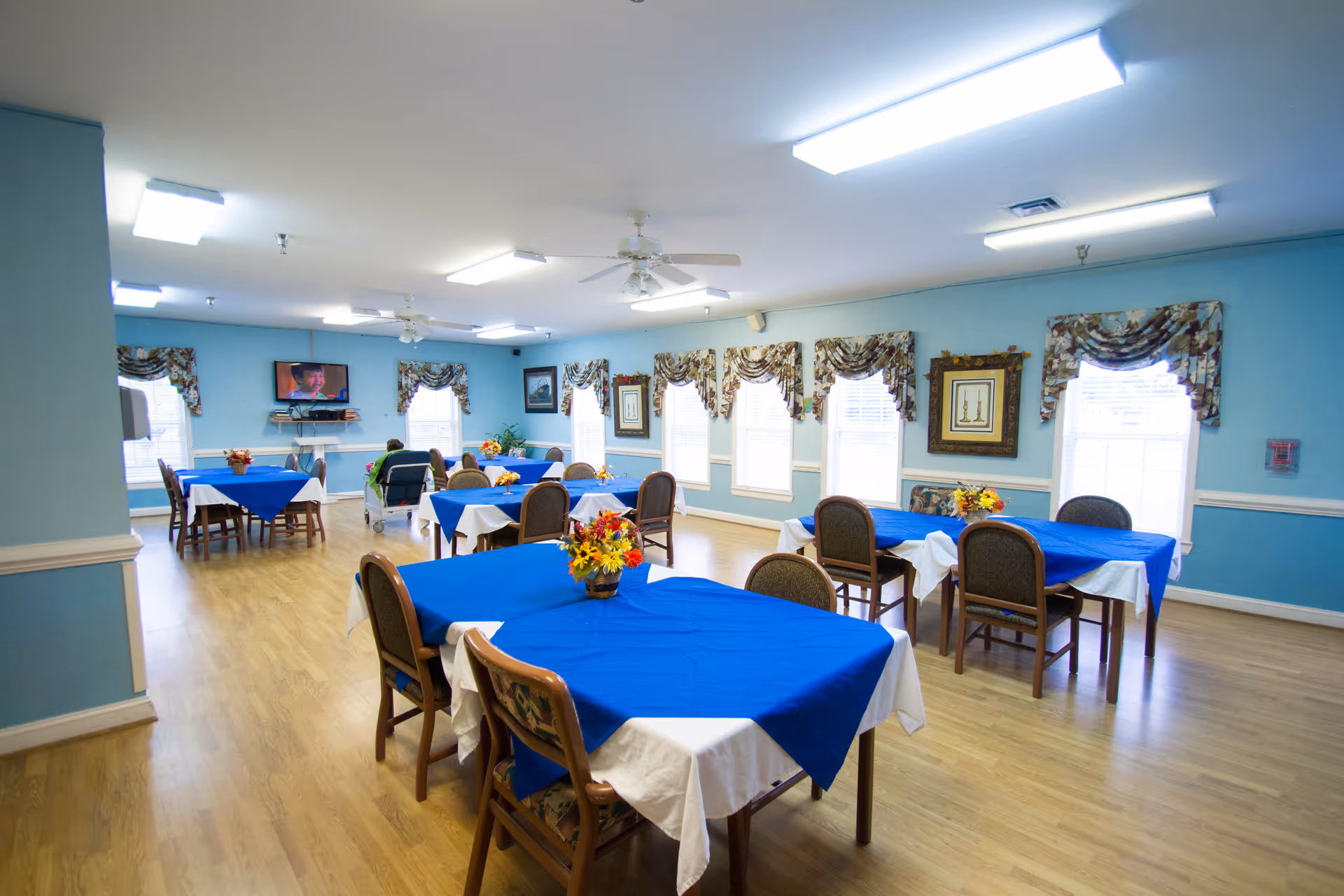 A bright dining room with several tables covered in white tablecloths and blue overlays, each decorated with a small flower arrangement. The room has light blue walls, multiple windows with floral valances, wooden flooring, and ceiling fans. A television is mounted on the far wall, and a few people are seated at the tables.