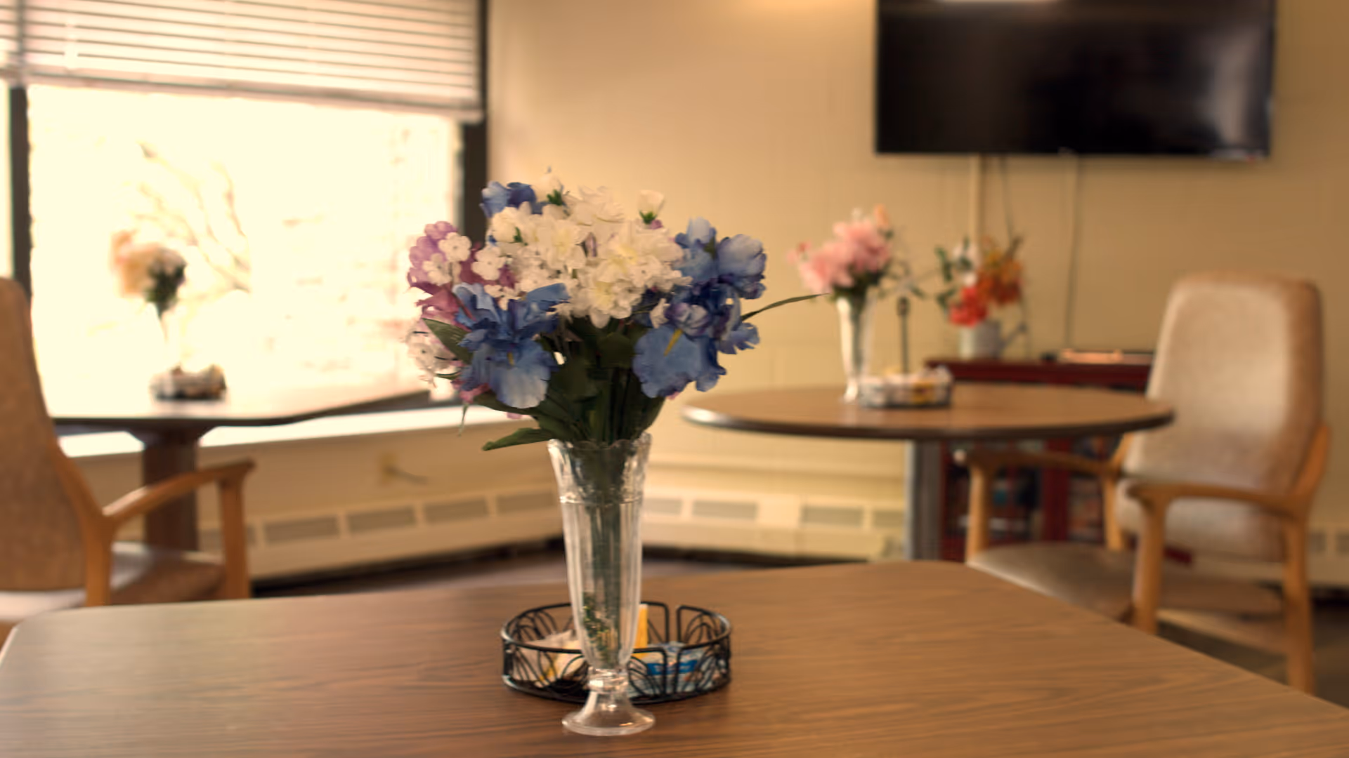 A cozy interior room with wooden tables and cushioned chairs. Each table has a vase with colorful artificial flowers. A large window with blinds lets in natural light, and a flat-screen TV is mounted on the wall in the background.
