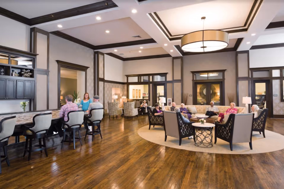 A spacious senior living common area with wooden floors and high ceilings featuring dark wood trim. Several seniors are seated in a circular arrangement on beige sofas and chairs around a central table with a decorative bowl. To the left, a few seniors sit on high chairs at a counter with a woman standing behind it. The room is well-lit with recessed lighting and a large hanging light fixture.