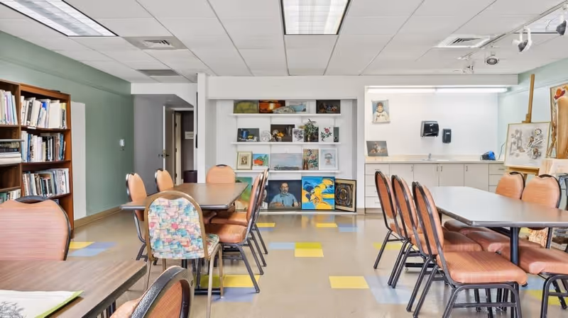 A well-lit art room with tables and chairs arranged for group activities. The room features shelves displaying various paintings and artworks, a cabinet with a sink, and a pastel-colored tiled floor with yellow and blue squares.