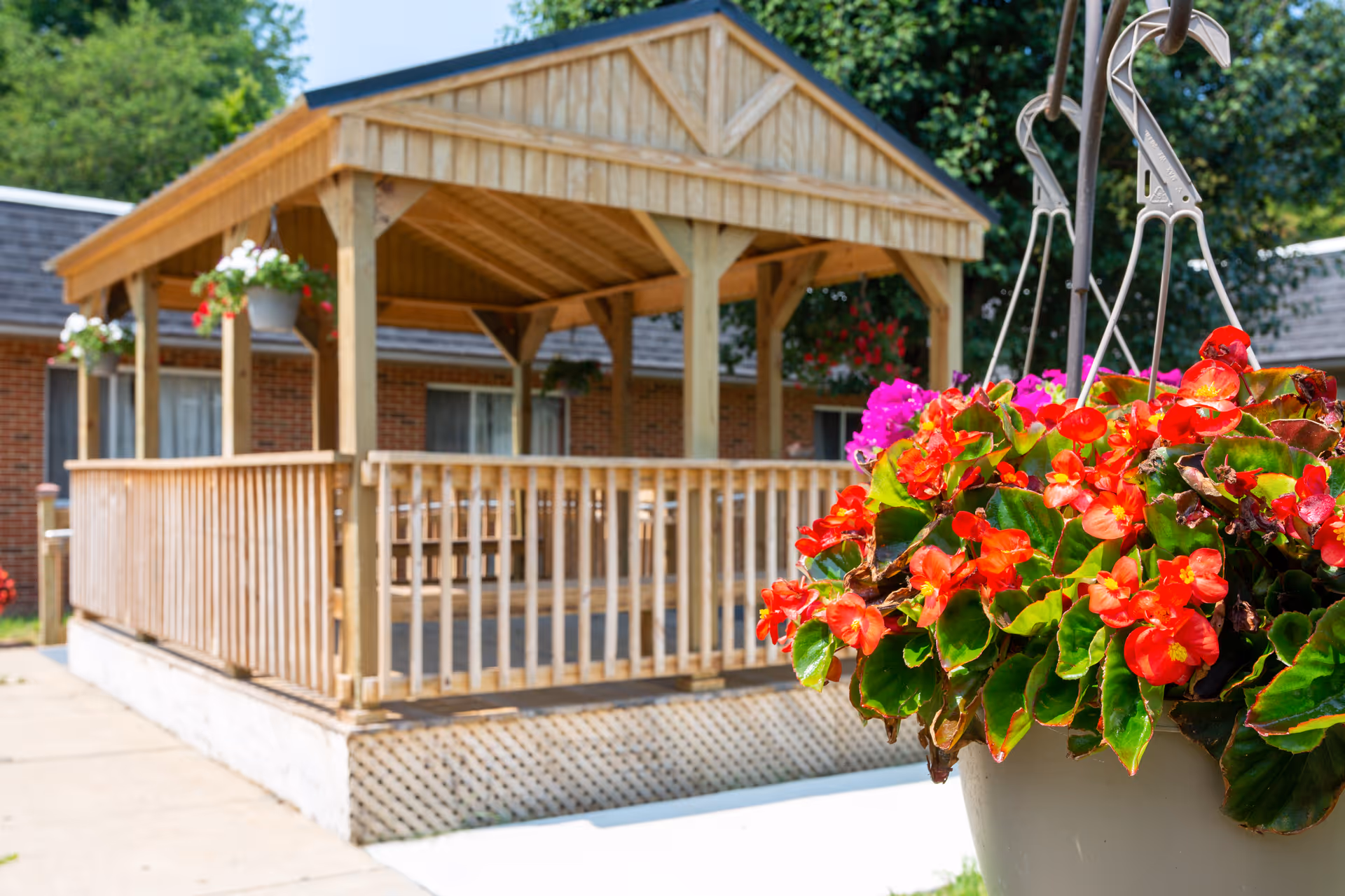 A wooden gazebo with a railing in an outdoor area of a senior living facility, with hanging flower pots containing red and pink flowers in the foreground and a brick building with windows in the background.