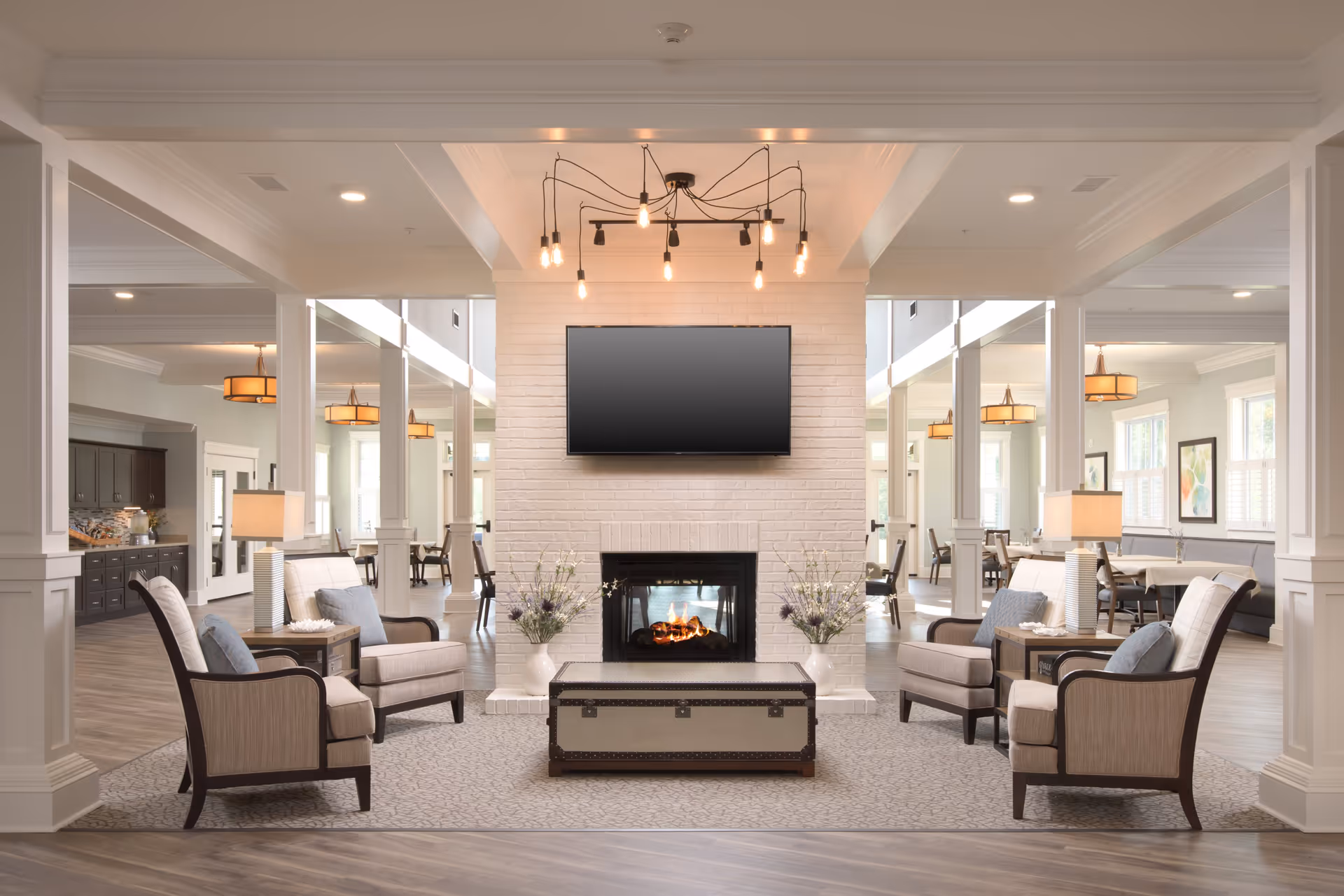 A bright and spacious senior living common area featuring a white brick fireplace with a mounted flat-screen TV above it. Four beige armchairs with blue cushions are arranged around a central trunk-style coffee table on a patterned rug. Two table lamps with beige shades sit on side tables beside the chairs. The room has large windows, modern light fixtures, and a view into a dining area with tables and chairs in the background.