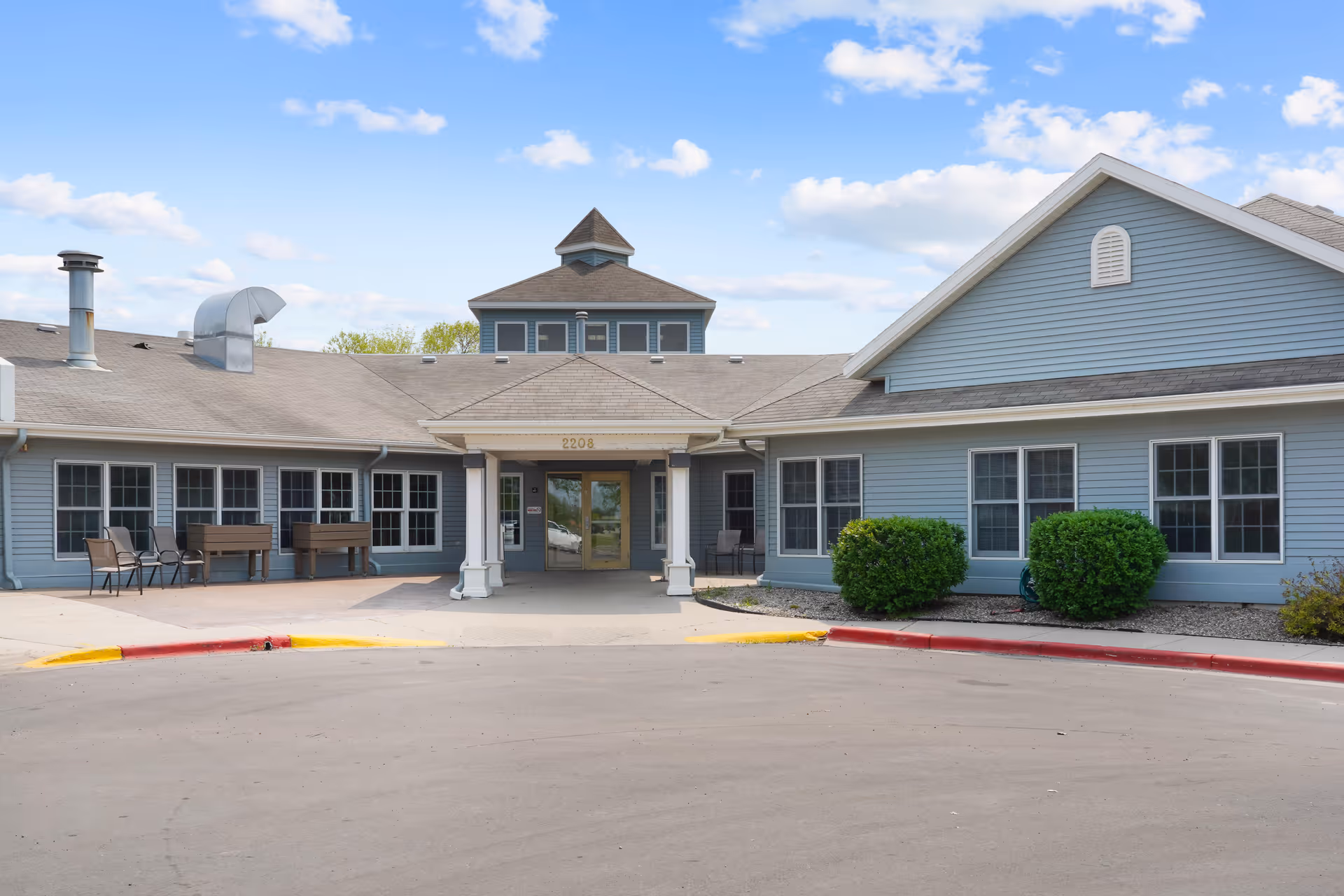 Front exterior of a gray single-story senior living building with a covered entrance, windows, outdoor seating and shrubs.