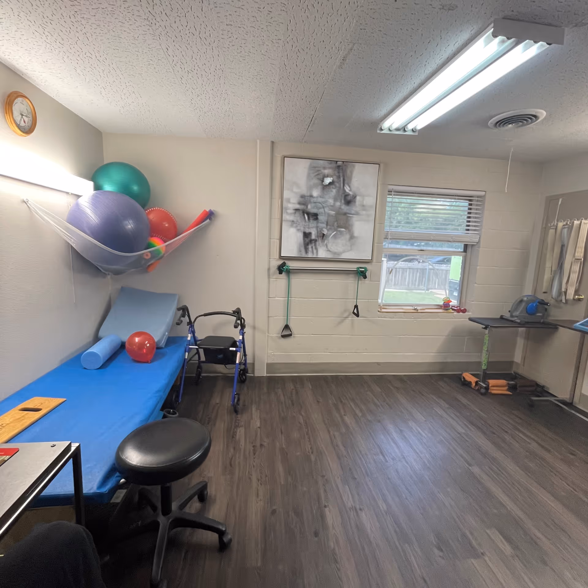 A physical therapy or rehabilitation room with exercise balls stored in a wall-mounted net, a blue padded therapy table with a red ball and foam roller on it, a walker, resistance bands hanging on the wall, a window with blinds, and a small table with exercise equipment. The room has wood-look flooring and fluorescent ceiling lights.