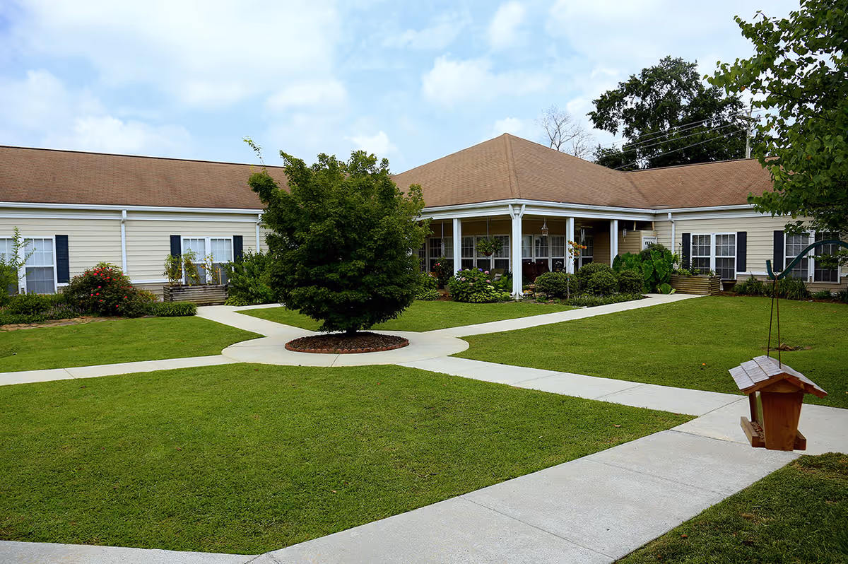 A well-maintained outdoor courtyard area of a senior living facility with green grass, a central tree surrounded by a circular mulch bed, intersecting concrete walkways, and a beige building with a brown roof and white columns in the background. There is a wooden bird feeder hanging on the right side.