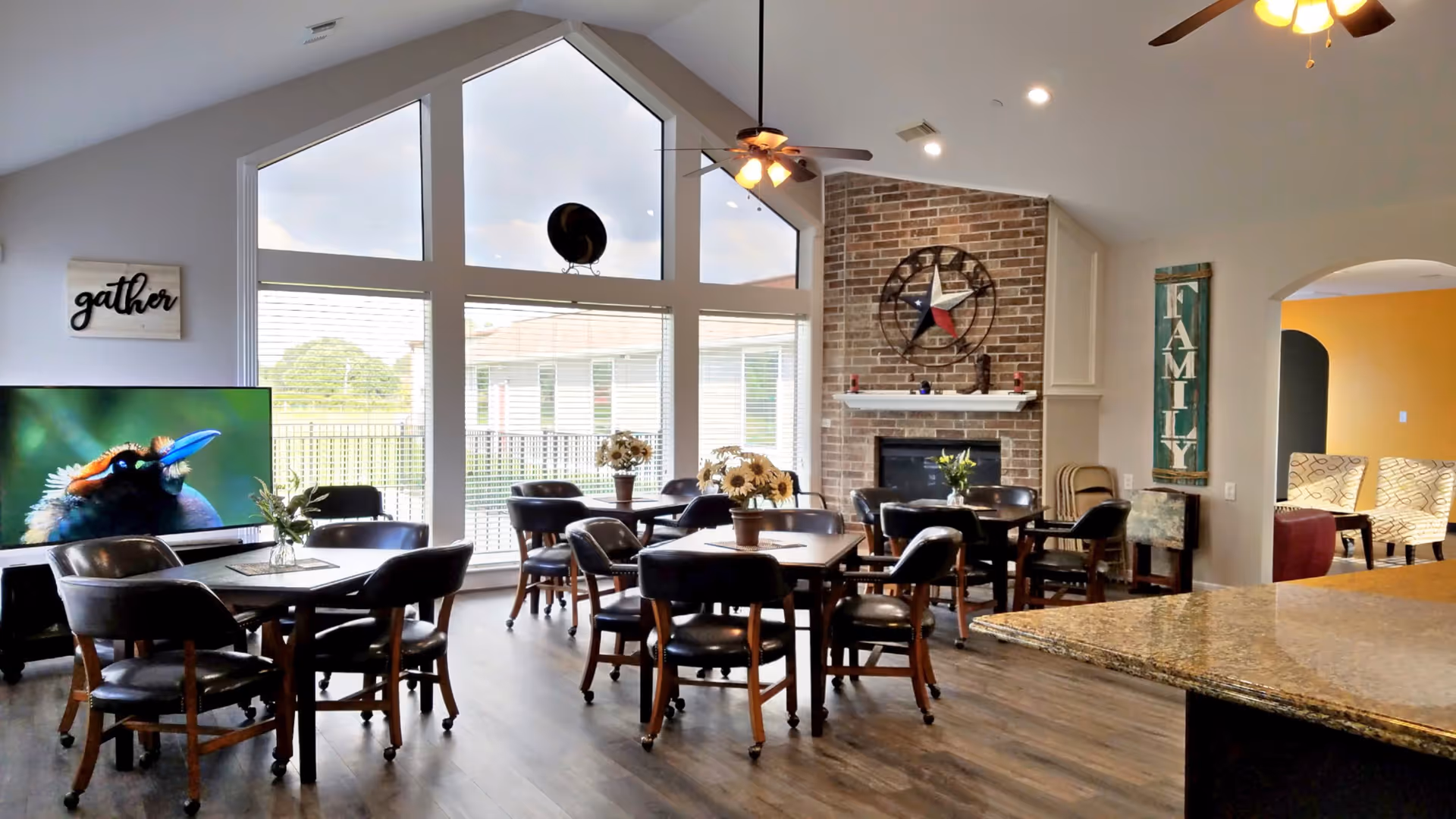 A bright and spacious common area with large windows letting in natural light. The room features several round tables with black chairs, a flat-screen TV displaying a bird, a brick fireplace with a decorative star above it, and wall decor including signs that say 'gather' and 'FAMILY'. The floor is wooden, and there is a granite countertop visible in the foreground.