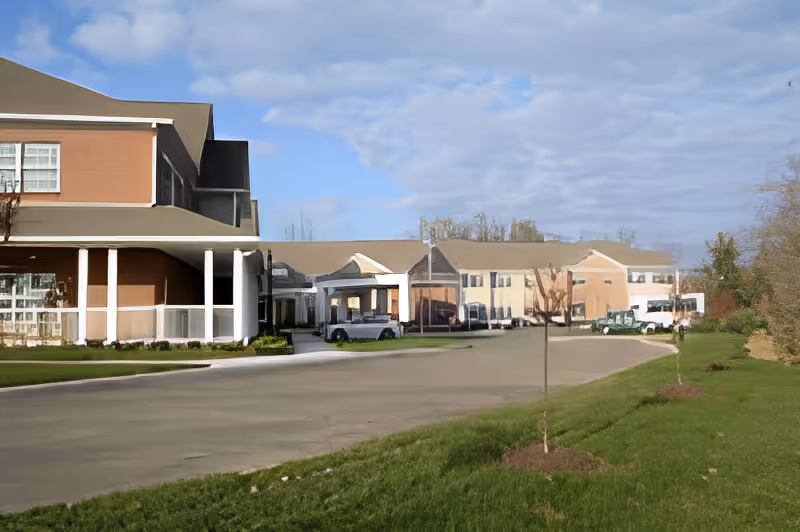 Exterior view of a senior living facility building with a driveway, green lawn, and small trees under a partly cloudy sky.