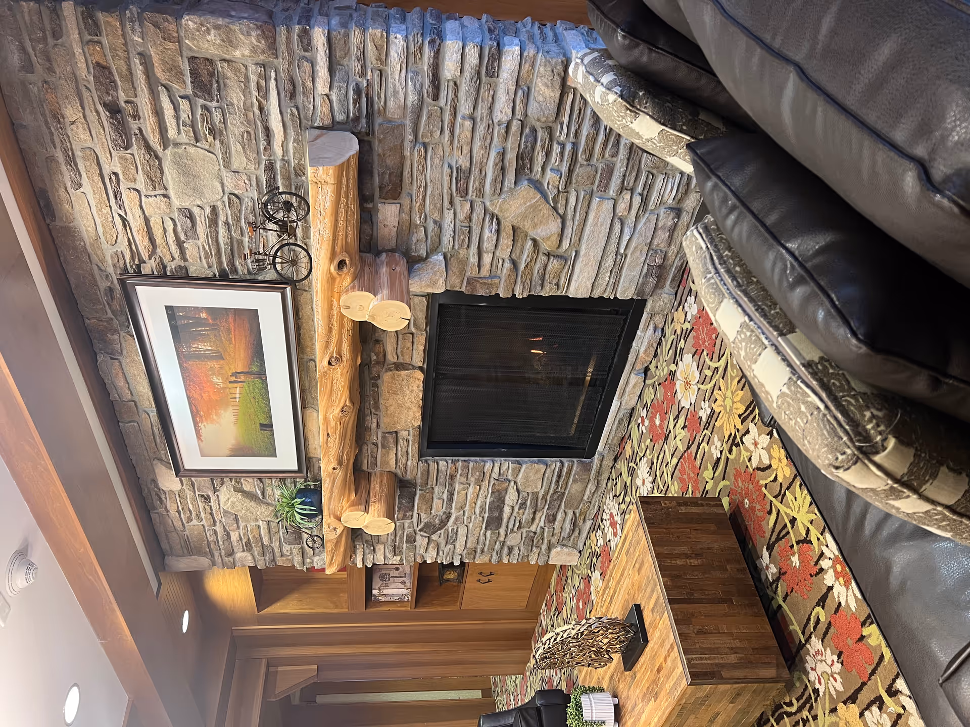 Cozy living room area featuring a stone fireplace with a wooden mantel decorated with a small plant and a miniature bicycle. Above the mantel hangs a framed picture of a scenic autumn landscape. The room has a patterned floral carpet, a wooden coffee table with decorative items, and a dark leather couch with patterned cushions.