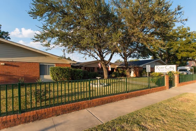 Exterior view of River City Care Center showing a single-story brick building with a green metal fence in front, a sidewalk, and several trees providing shade on a sunny day.