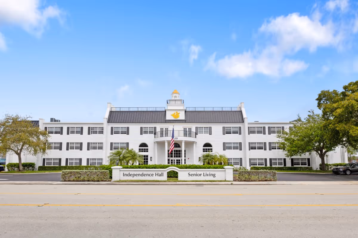 Front view of a white three-story senior living building with a central portico, American flag, and a sign reading 'Independence Hall Senior Living'.