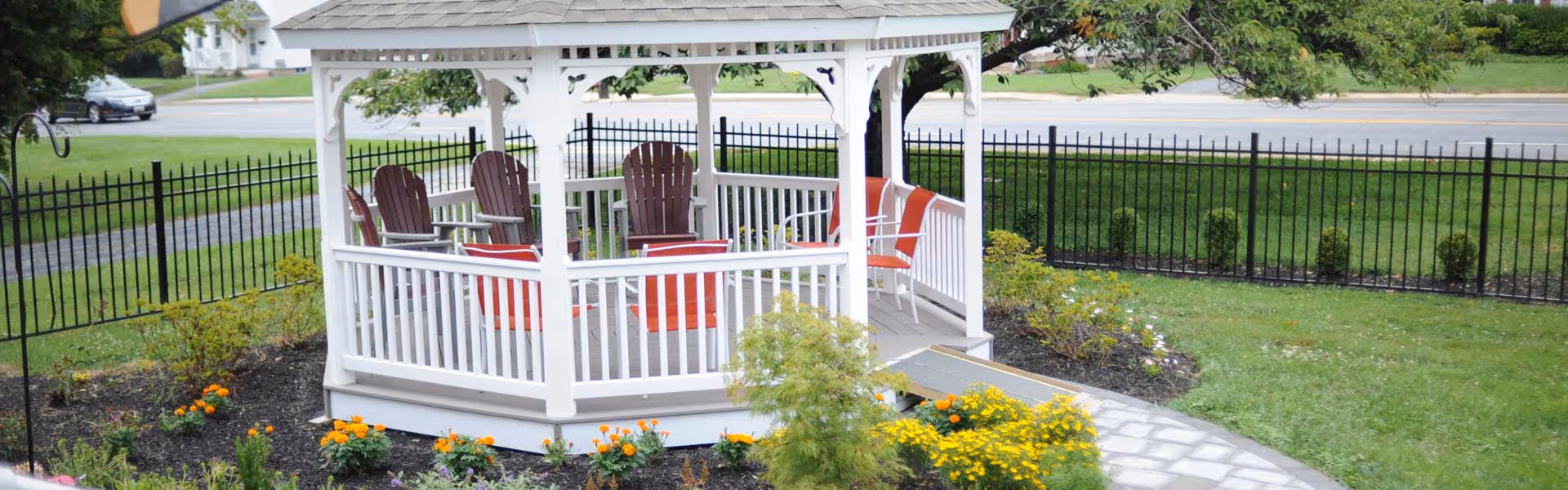 A white wooden gazebo with a shingled roof situated in a garden area. Inside the gazebo are several red Adirondack chairs arranged around a small table. The gazebo is surrounded by green grass, flower beds with yellow and orange flowers, and a black metal fence enclosing the area. A paved walkway leads up to the gazebo.