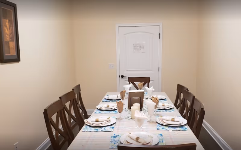 Long dining table set for eight with place settings and a centerpiece in a small beige dining room with a white door at the far end.