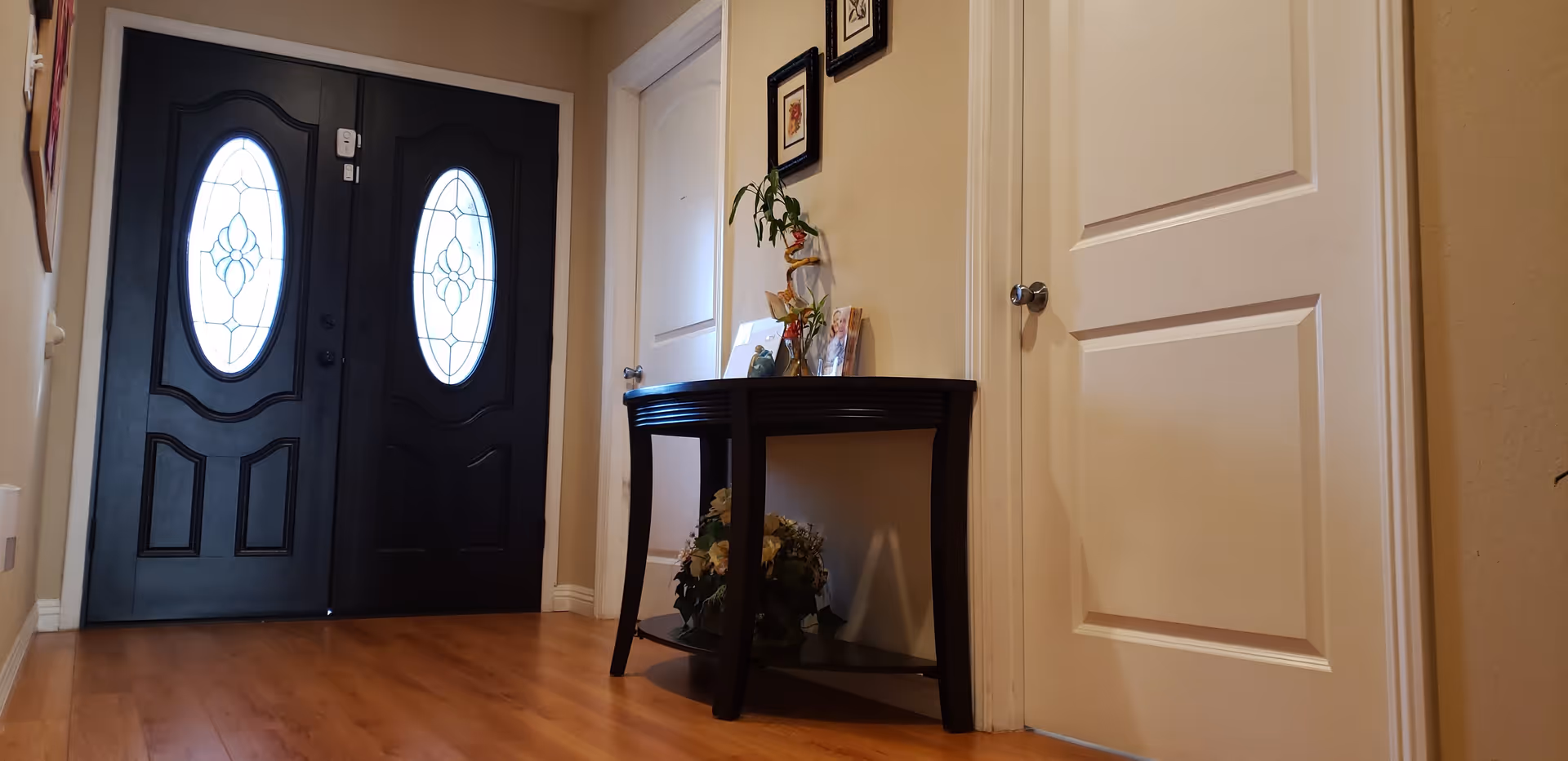 Interior view of a hallway with a black double front door featuring oval glass panels. There is a small dark wooden table against the wall with decorative items including a plant, framed pictures, and a flower arrangement underneath. Two white doors are visible on the right side of the hallway.