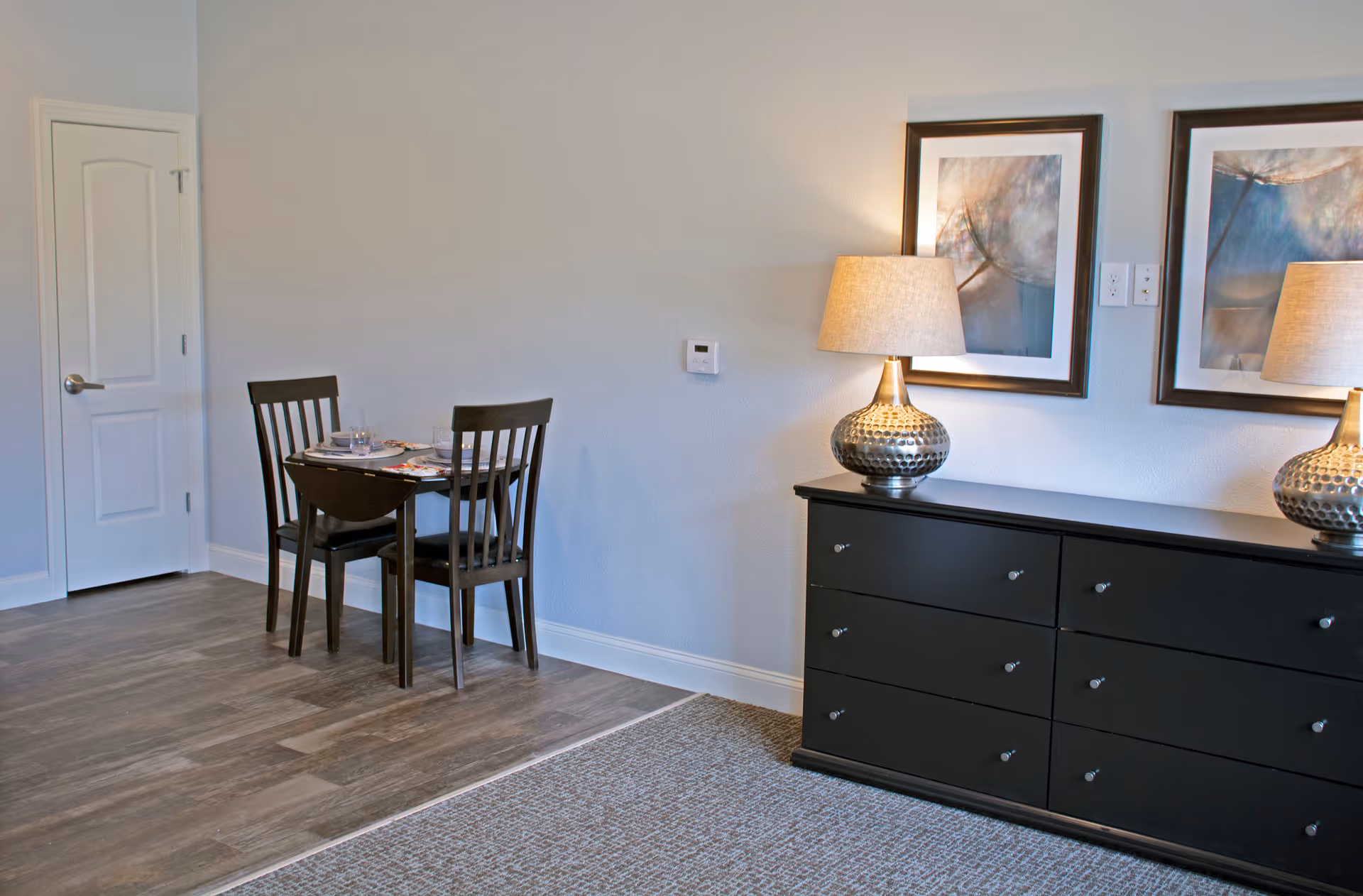 A small dining area with a dark wooden table set for two with plates and glasses, next to a black dresser with six drawers. On top of the dresser are two matching lamps with beige shades and two framed abstract artworks hanging on the wall above it. The floor is a combination of wood and carpet, and there is a white door in the background.