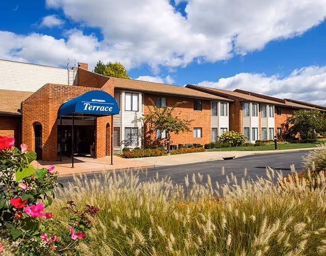 Exterior view of Bethesda Terrace senior living facility showing a two-story brick building with a blue awning labeled 'Terrace' over the entrance. The foreground features ornamental grasses and flowering plants under a partly cloudy sky.