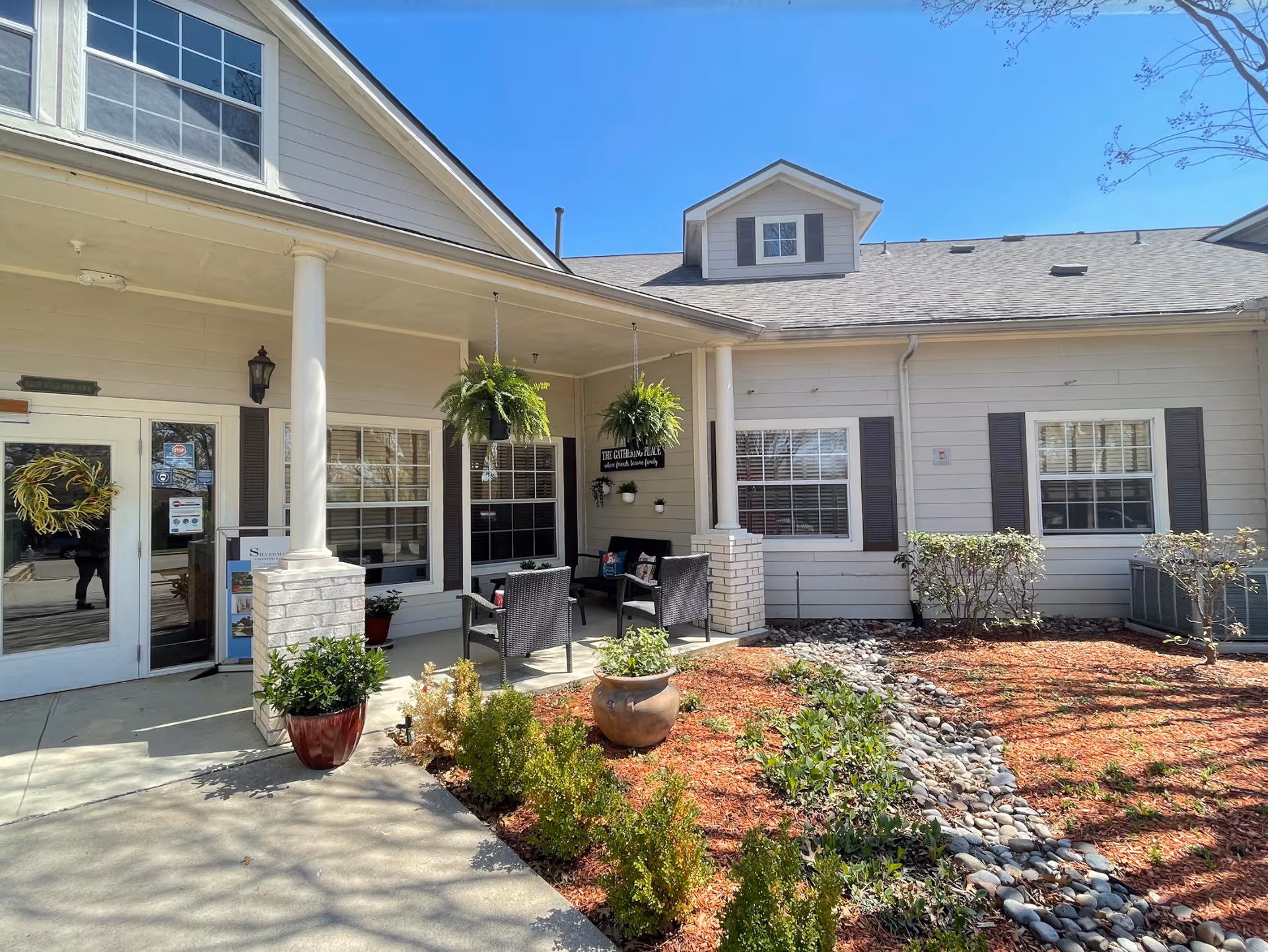 Exterior view of a senior living facility entrance with a covered porch featuring white columns, hanging plants, outdoor seating with chairs and a small table, and landscaped garden beds with shrubs and rocks under a clear blue sky.