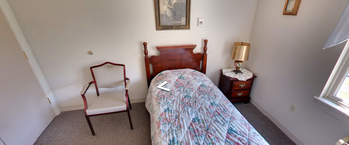 A small bedroom with a single wooden bed covered with a floral quilt. Next to the bed is a wooden nightstand with a lace doily and a gold-colored lamp. There is a cushioned armchair with wooden arms and legs positioned against the wall. A framed picture hangs above the bed, and a window on the right side lets in natural light.